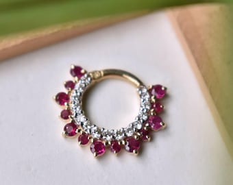 Puede incluir: Un anillo de septum dorado con un diseño circular. El anillo está adornado con pequeñas piedras preciosas redondas de color rojo rubí y cristales transparentes. Las piedras preciosas están dispuestas alrededor del borde exterior y el círculo interior.
