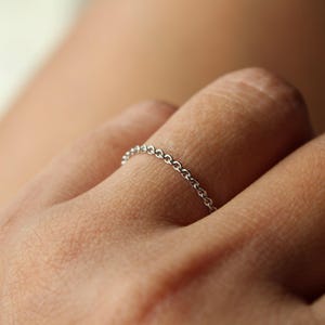 May include: A delicate silver chain ring. The ring is composed of small, interconnected links, forming a continuous chain. The ring is worn on a finger, showcasing its minimalist design and subtle elegance. The ring's simple design makes it suitable for everyday wear.