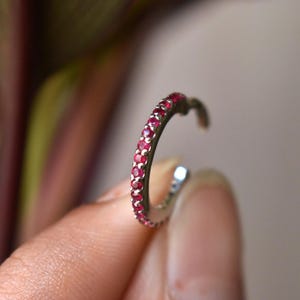 May include: A silver-toned ear cuff adorned with a row of small, vibrant red gemstones. The curved design is held by a person's fingers, showcasing the jewelry's delicate details and craftsmanship. The gemstones are evenly spaced along the cuff.
