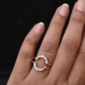 May include: A gold-colored ring with a half-circle design, adorned with small, sparkling white gemstones. The ring is held by a hand against a dark background, showcasing the ring's intricate details and elegant style. The ring is a jewelry item.