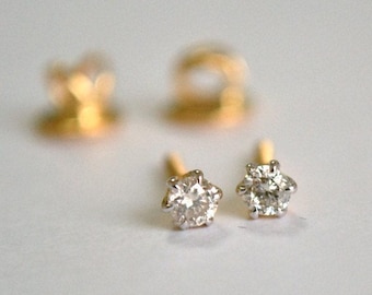 Puede incluir: Un par de pendientes de diamantes con seis garras, engastados en un poste de oro. Los diamantes son redondos y claros, reflejando la luz. Dos cierres dorados para pendientes están ligeramente desenfocados en el fondo.