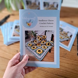 May include: A light blue booklet with the text "Sunflower Throw Crochet Pattern" and a photo of a crocheted sunflower blanket. The booklet has a floral logo that says "libbycraft". Other booklets are in the background.