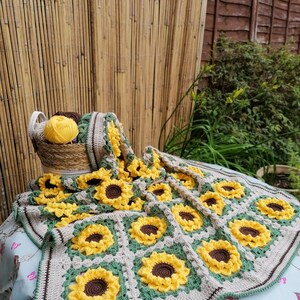 Puede incluir: Una manta de ganchillo con forma de girasol con un borde marr&oacute;n y verde. La manta est&aacute; hecha de hilo beige, amarillo y verde. La manta est&aacute; colocada sobre un mantel con estampado azul y blanco.
