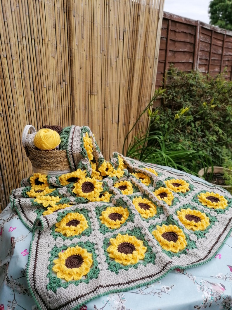 Puede incluir: Una manta de ganchillo con forma de girasol con un borde marr&oacute;n y verde. La manta est&aacute; hecha de hilo beige, amarillo y verde. La manta est&aacute; colocada sobre un mantel con estampado azul y blanco.