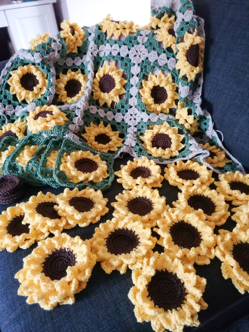 Puede incluir: Una manta de ganchillo con un dise&ntilde;o de girasol. La manta est&aacute; hecha de hilo verde, marr&oacute;n y beige. Hay muchos motivos de girasol individuales en primer plano.