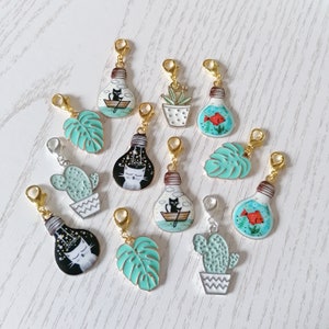 May include: A set of 10 colorful charm pendants featuring various designs, including a cat in a lightbulb, a fish in a bowl, a cactus, and a monstera leaf. The charms are made of metal and have a gold finish.