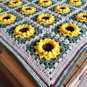 Puede incluir: Una manta de ganchillo con un patr&oacute;n de girasol en tonos de amarillo, verde, marr&oacute;n y beige. La manta est&aacute; hecha con un punto de granny square y tiene un borde en marr&oacute;n y verde.