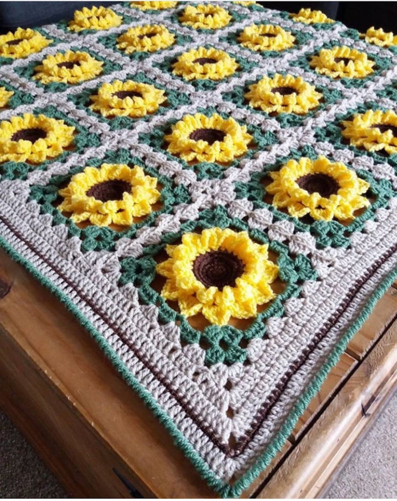 Puede incluir: Una manta de ganchillo con un patr&oacute;n de girasol en tonos de amarillo, verde, marr&oacute;n y beige. La manta est&aacute; hecha con un punto de granny square y tiene un borde en marr&oacute;n y verde.