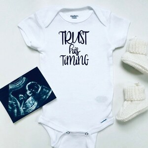 May include: White baby onesie with the text "TRUST his TIMING" in black script. A black and white ultrasound image and a pair of white knitted baby booties are on a white surface.