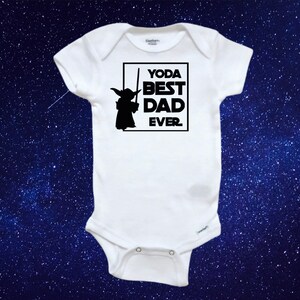 May include: White baby onesie with the text "YODA BEST DAD EVER" in a black square, along with a black silhouette of Yoda holding a lightsaber. The onesie has short sleeves and snap closures. The background is a dark blue with white stars.