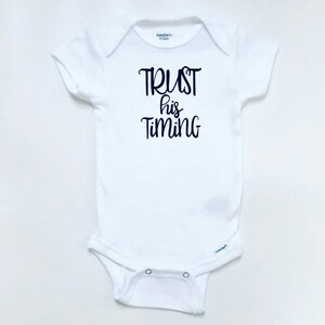 May include: White baby onesie with black text that reads "TRUST his TIMING".
