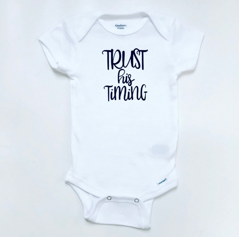 May include: White baby onesie with black text that reads "TRUST his TIMING".