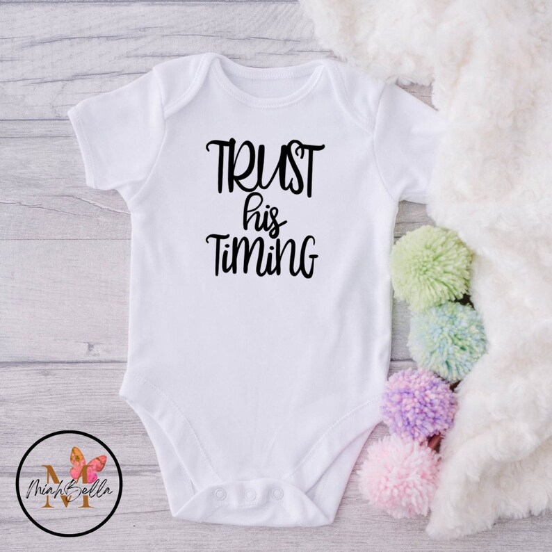 May include: White baby onesie with black text that reads "TRUST his TIMING".