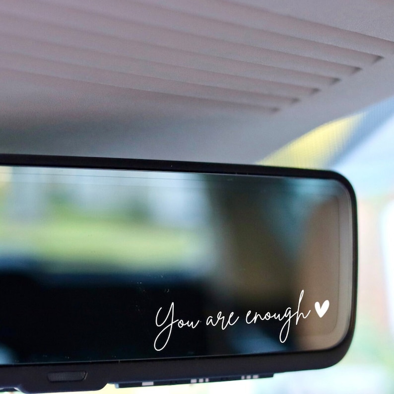 K&ouml;nnte beinhalten: Ein R&uuml;ckspiegel im Auto mit dem Schriftzug "You are enough" in zarter, wei&szlig;er Schreibschrift, erg&auml;nzt durch ein kleines Herzsymbol. Der Text hebt sich vom dunklen Spiegelhintergrund ab.