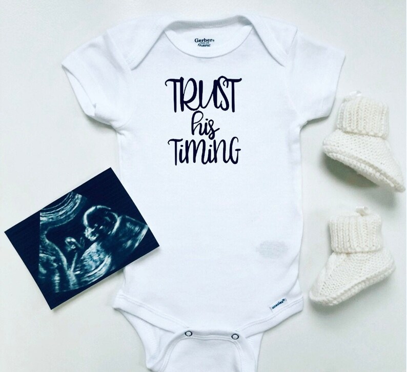 May include: White baby onesie with the text "TRUST his TIMING" in black script. A black and white ultrasound image and a pair of white knitted baby booties are on a white surface.