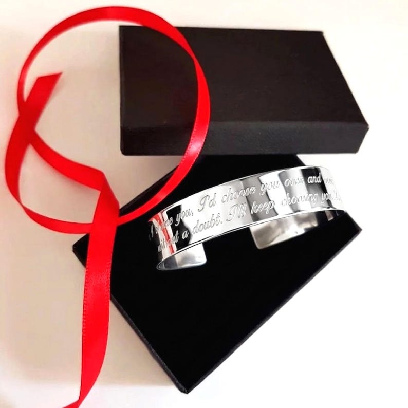 May include: A silver cuff bracelet with engraved text, presented in a black gift box with a red ribbon. The bracelet has the inscription: "I choose you over and without a doubt. I'll keep choosing you."
