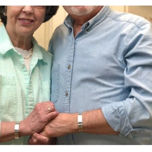 May include: Two people wearing matching silver metal cuff bracelets. The bracelets have a simple, rectangular design. The person on the left is wearing a light green shirt, and the person on the right is wearing a light blue shirt.