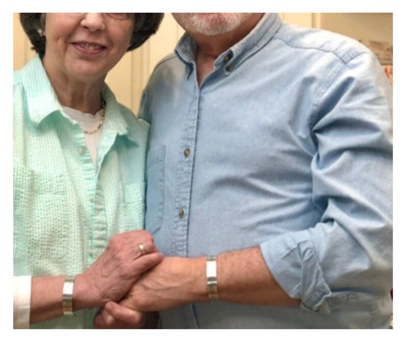 May include: Two people wearing matching silver metal cuff bracelets. The bracelets have a simple, rectangular design. The person on the left is wearing a light green shirt, and the person on the right is wearing a light blue shirt.