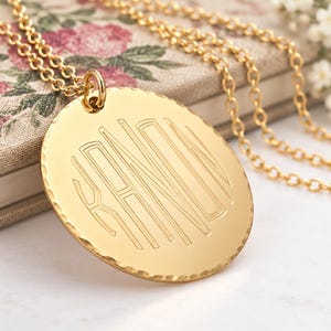 Könnte beinhalten: Eine goldfarbene, kreisförmige Anhängerkette mit gehämmertem Rand und einem mittig eingravierten Monogramm mit den Buchstaben K, A, N, O, N. Der Anhänger hängt an einer zarten goldfarbenen Kette mit polierter Oberfläche und feinen Linien.