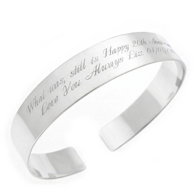 May include: A silver cuff bracelet with engraved text. The text reads "What was, still is Happy 20th Anniversary Love You Always Liz. 04/03/04". The bracelet has a polished finish and an open design.