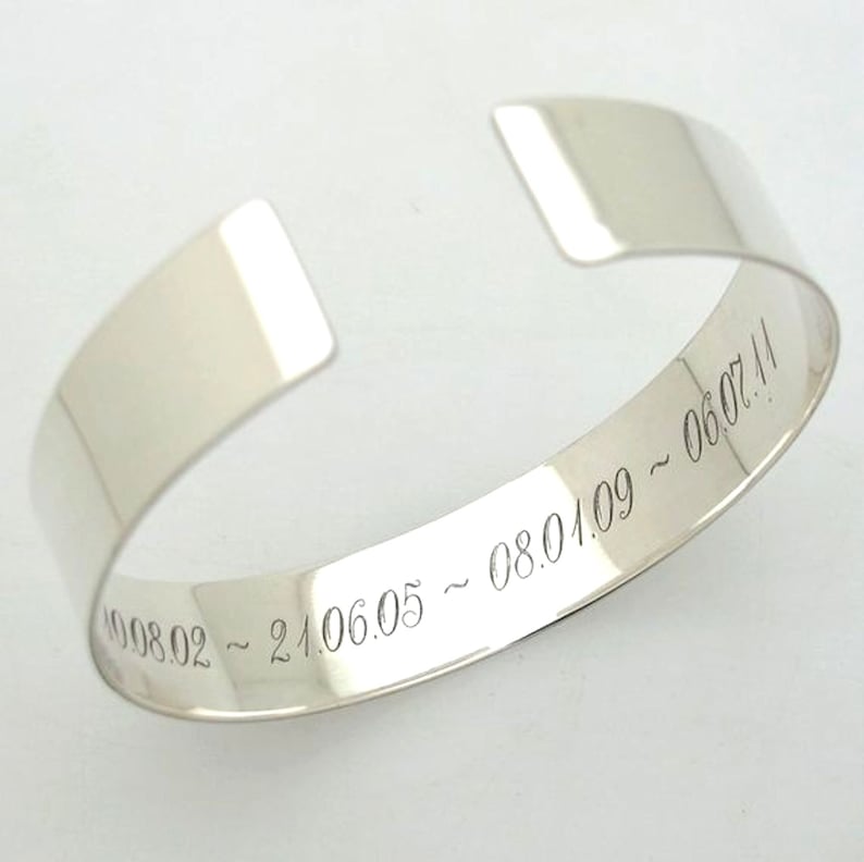 May include: A silver cuff bracelet with engraved dates. The open-ended bracelet has a smooth, polished finish. The dates are engraved in a delicate, cursive font. The bracelet is a simple, elegant design.