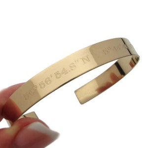 May include: Gold-toned metal bracelet with the coordinates "40° 56' 54.8" N" engraved on the band.