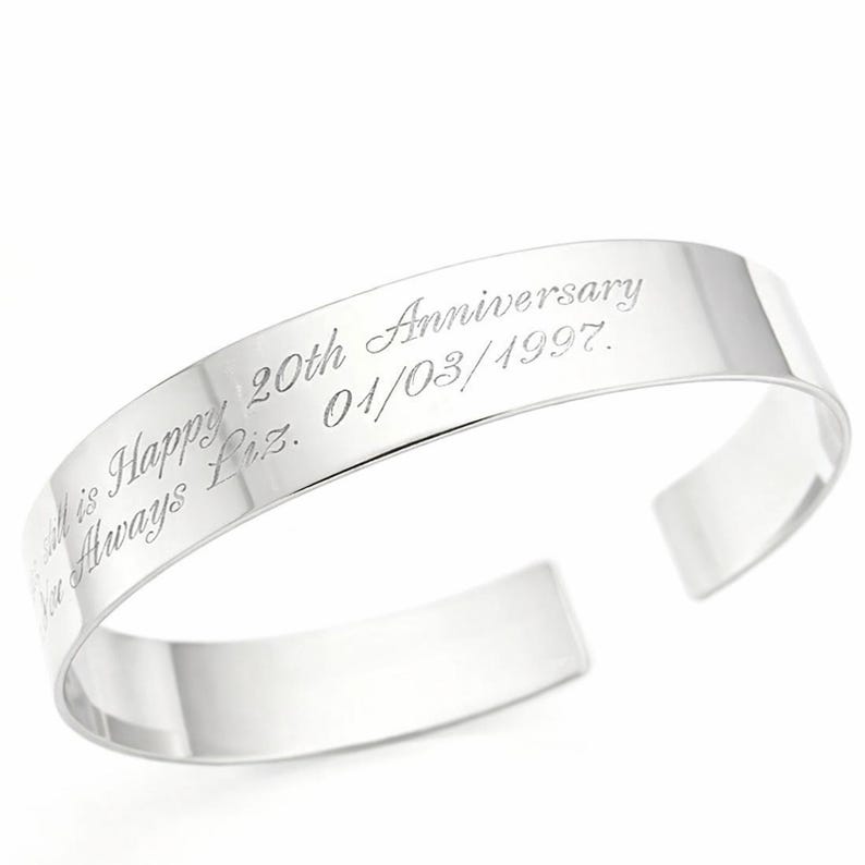 May include: A polished silver cuff bracelet with engraved text. The text reads "All is Happy 20th Anniversary I'm Always Liz. 01/03/1997." The bracelet has a simple, open design.