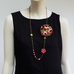 May include: A long gold chain necklace with green, pink, and red beads. A large round floral brooch with red, pink, and green embroidery is attached to the chain.