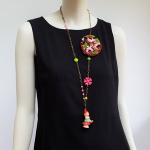 May include: A long necklace with a round floral embroidered pendant, pink and green beads, a pink flower charm, and a small gnome figurine charm.