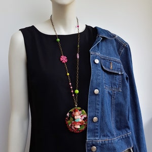May include: A gold chain necklace with colorful beads and a round pendant featuring a miniature doll wearing a red dress and a green hat. The pendant is surrounded by red, green, and black beads.