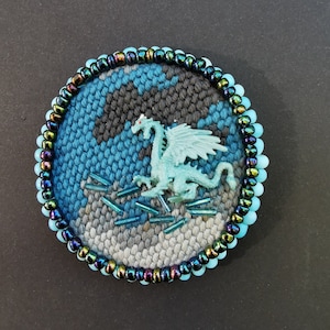 Peut inclure: Une tapisserie murale ronde tissée avec un motif bleu, gris et noir. Un petit dragon en plastique bleu clair se trouve au centre du motif. La tapisserie est entourée d'une bordure de perles bleues irisées.