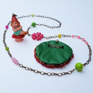 May include: A green beaded necklace with a pink flower charm and a gnome figurine charm. The necklace is made with a brown chain and has pink, green, and yellow beads.
