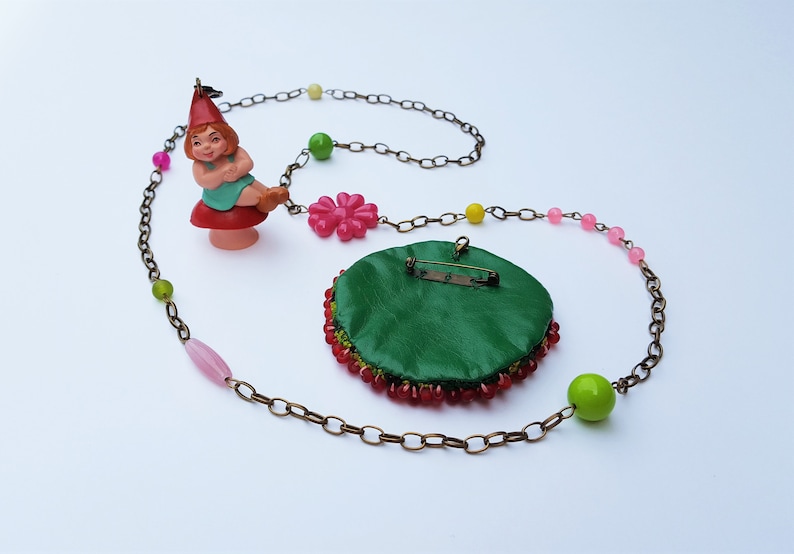 May include: A green beaded necklace with a pink flower charm and a gnome figurine charm. The necklace is made with a brown chain and has pink, green, and yellow beads.