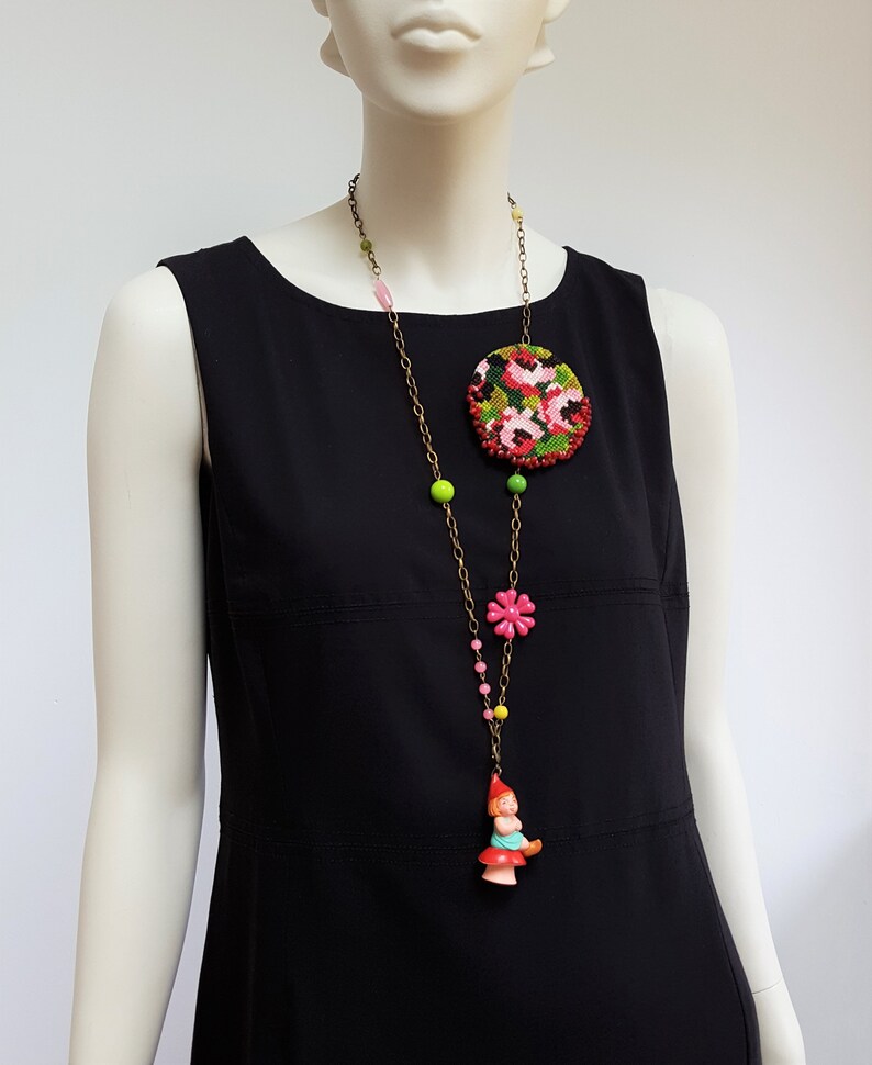 May include: A long necklace with a round floral embroidered pendant, pink and green beads, a pink flower charm, and a small gnome figurine charm.