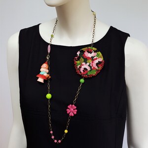 May include: A long gold chain necklace with pink, green, and yellow beads. The necklace features a large round floral embroidered pendant and a small plastic figurine of a girl sitting on a mushroom.