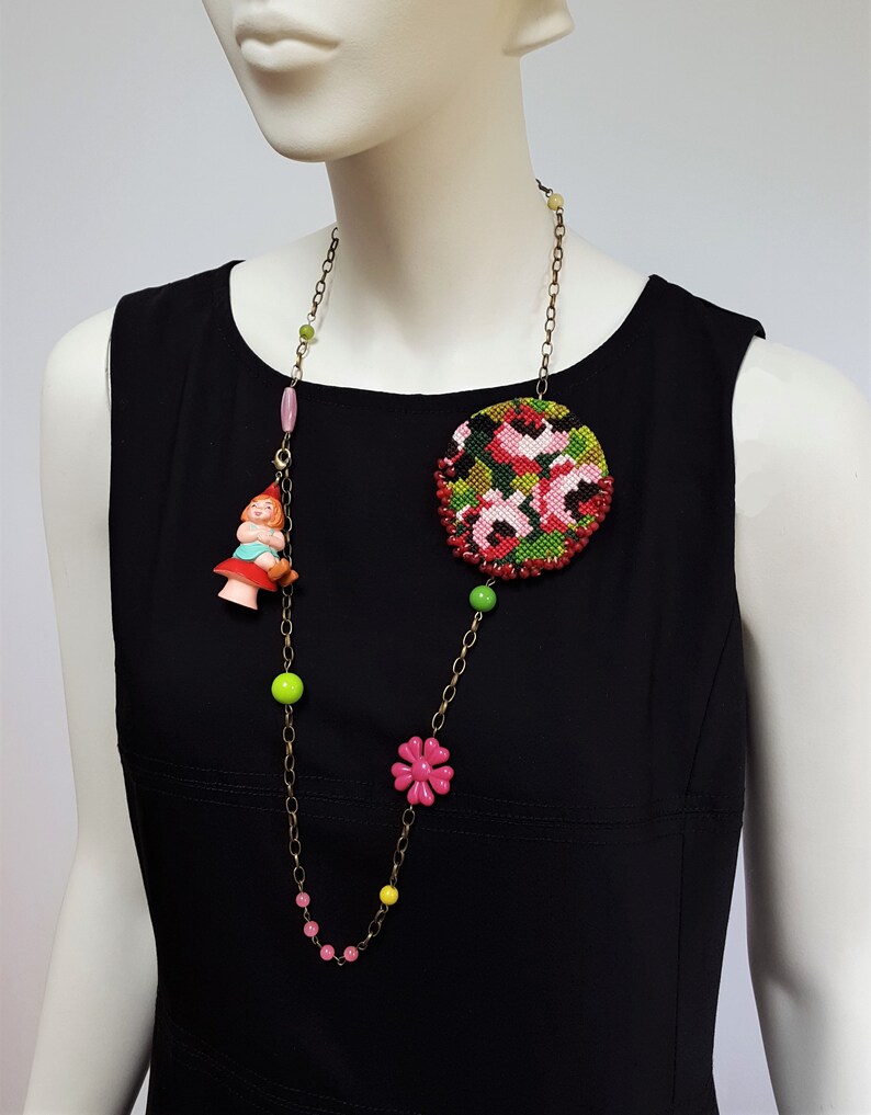 May include: A long gold chain necklace with pink, green, and yellow beads. The necklace features a large round floral embroidered pendant and a small plastic figurine of a girl sitting on a mushroom.