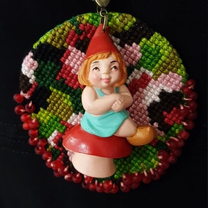 May include: A red and green embroidered circle with a red beaded border. A small plastic figurine of a girl wearing a red hat and blue dress sits on a red mushroom in the center of the circle.