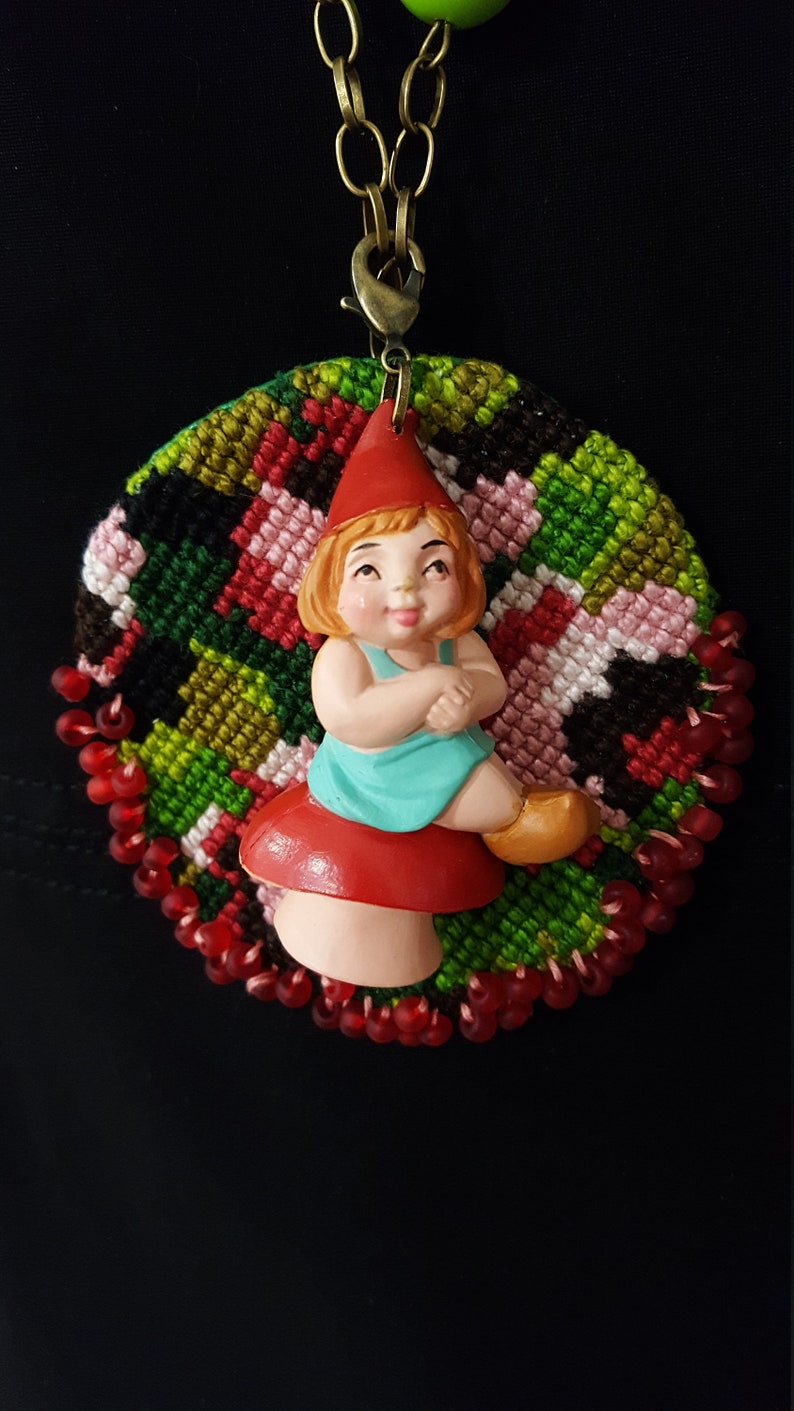 May include: A red and green embroidered circle with a red beaded border. A small plastic figurine of a girl wearing a red hat and blue dress sits on a red mushroom in the center of the circle.
