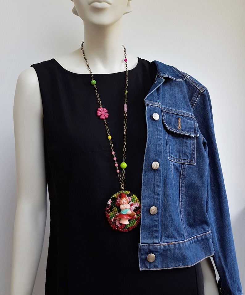 May include: A gold chain necklace with colorful beads and a round pendant featuring a miniature doll wearing a red dress and a green hat. The pendant is surrounded by red, green, and black beads.