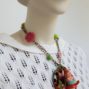 May include: A colorful necklace with a green, pink, and red embroidered oval pendant featuring a small gnome figurine. The necklace also has green, yellow, and pink beads.