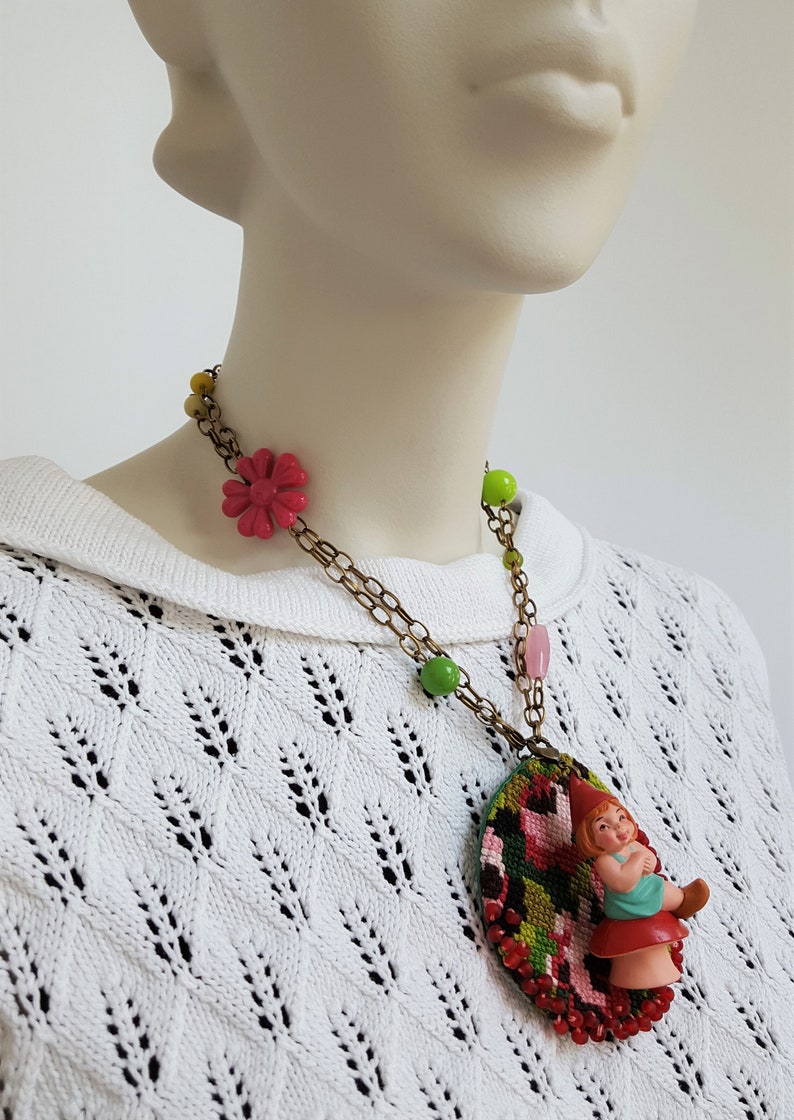 May include: A colorful necklace with a green, pink, and red embroidered oval pendant featuring a small gnome figurine. The necklace also has green, yellow, and pink beads.