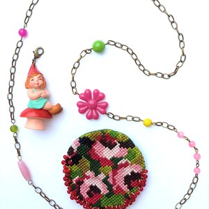 May include: A gold chain necklace with pink, green, and yellow beads. The necklace has a pink flower charm, a gnome charm, and a round embroidered charm with pink flowers on a green background.
