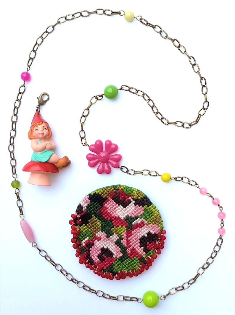 May include: A gold chain necklace with pink, green, and yellow beads. The necklace has a pink flower charm, a gnome charm, and a round embroidered charm with pink flowers on a green background.