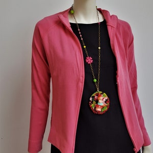 May include: A pink zip-up jacket with a long, colorful beaded necklace featuring a round pendant with a small doll figure. The pendant is made of beads and has a red, green, and black pattern.