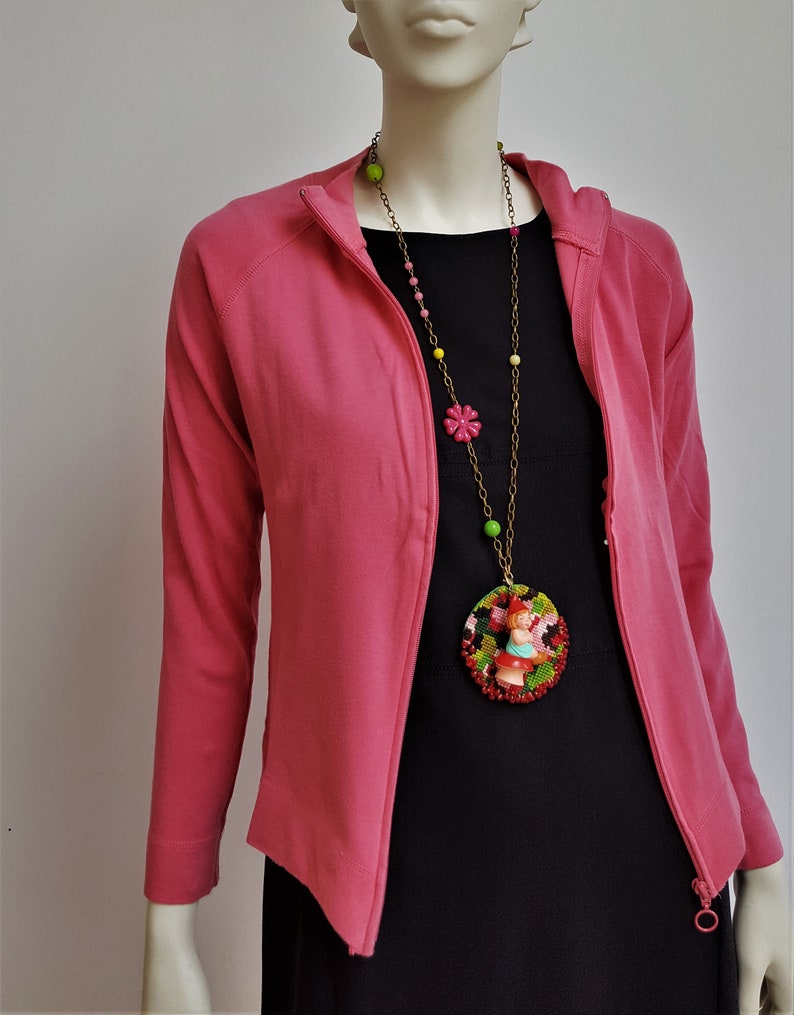 May include: A pink zip-up jacket with a long, colorful beaded necklace featuring a round pendant with a small doll figure. The pendant is made of beads and has a red, green, and black pattern.