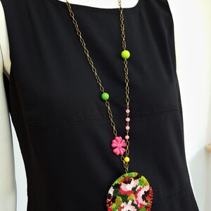 May include: A long gold chain necklace with pink, green, and yellow beads. The necklace has a large round pendant with a floral design in shades of red, pink, green, and black.