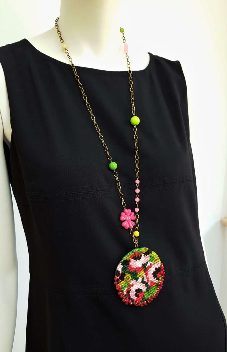 May include: A long gold chain necklace with pink, green, and yellow beads. The necklace has a large round pendant with a floral design in shades of red, pink, green, and black.