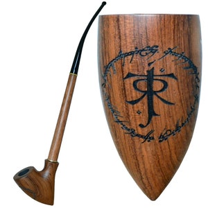 Puede incluir: Una pipa de madera con una larga boquilla curva y una cazoleta tallada con un diseño. La pipa está junto a un soporte de madera con la inscripción "Un anillo para gobernarlos a todos" en escritura élfica.