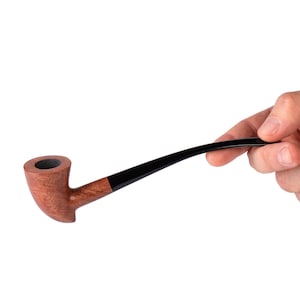 Puede incluir: Una pipa de tabaco de madera con una boquilla negra curvada. El cuenco de la pipa es de color marrón rojizo, y la boquilla es sostenida por una mano sobre un fondo blanco. La pipa está diseñada para fumar tabaco.