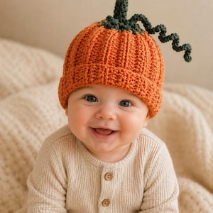 May include: A smiling baby wearing an orange crochet pumpkin hat with a green stem and curly vine. The baby is dressed in a beige waffle-knit onesie with wooden buttons. The background is a soft, neutral color.